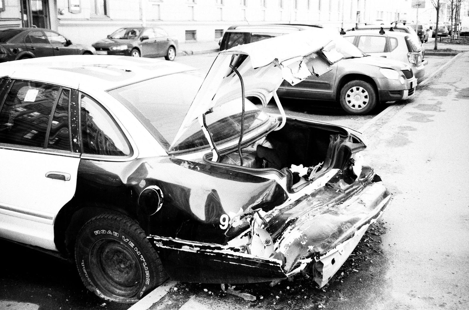 A black and white photo of a wrecked car with heavy damage on an urban street.