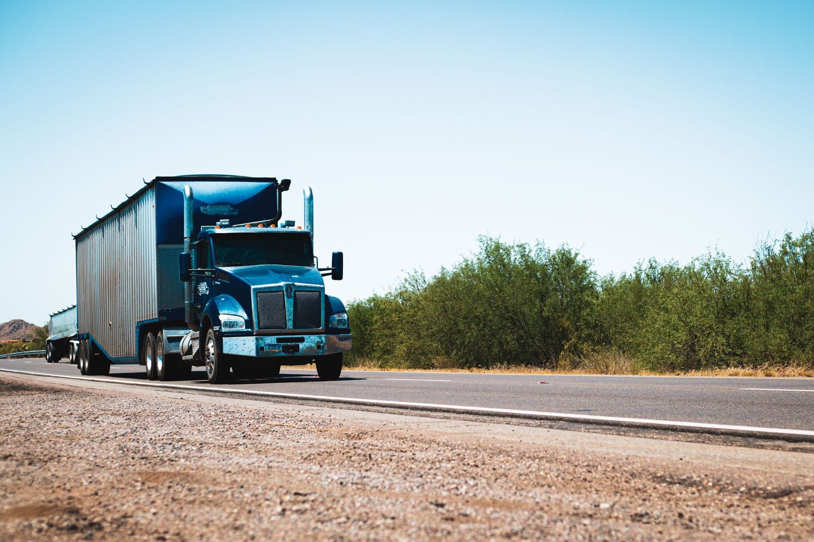 A blue semi truck drives down a rural highway under a clear blue sky.
