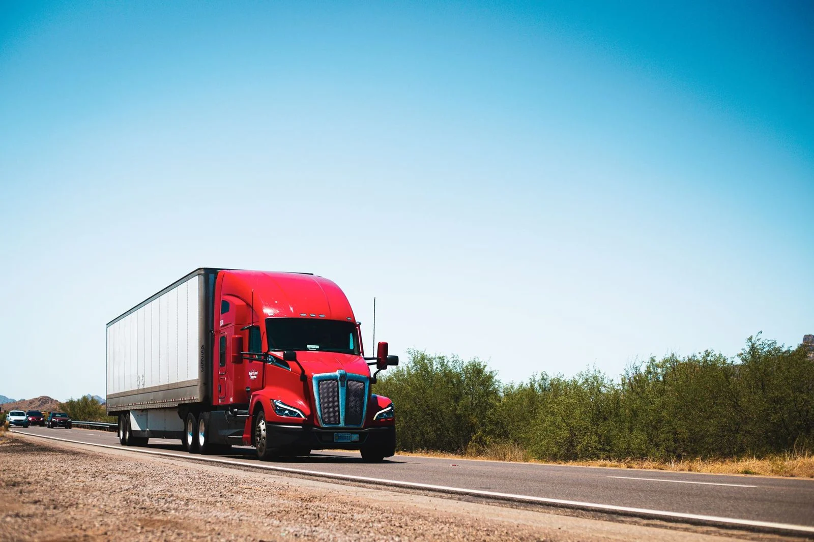 Red semi-trailer truck driving on an open highway under a clear blue sky.
