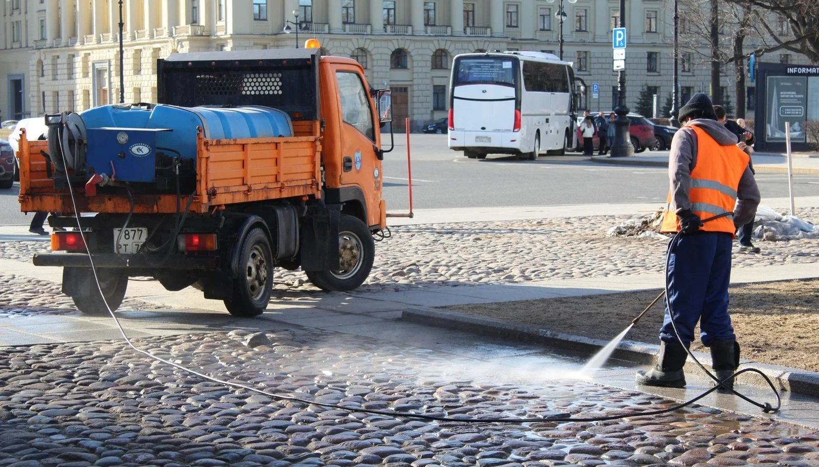 A city worker sprays a water hose across a sunlit urban street.