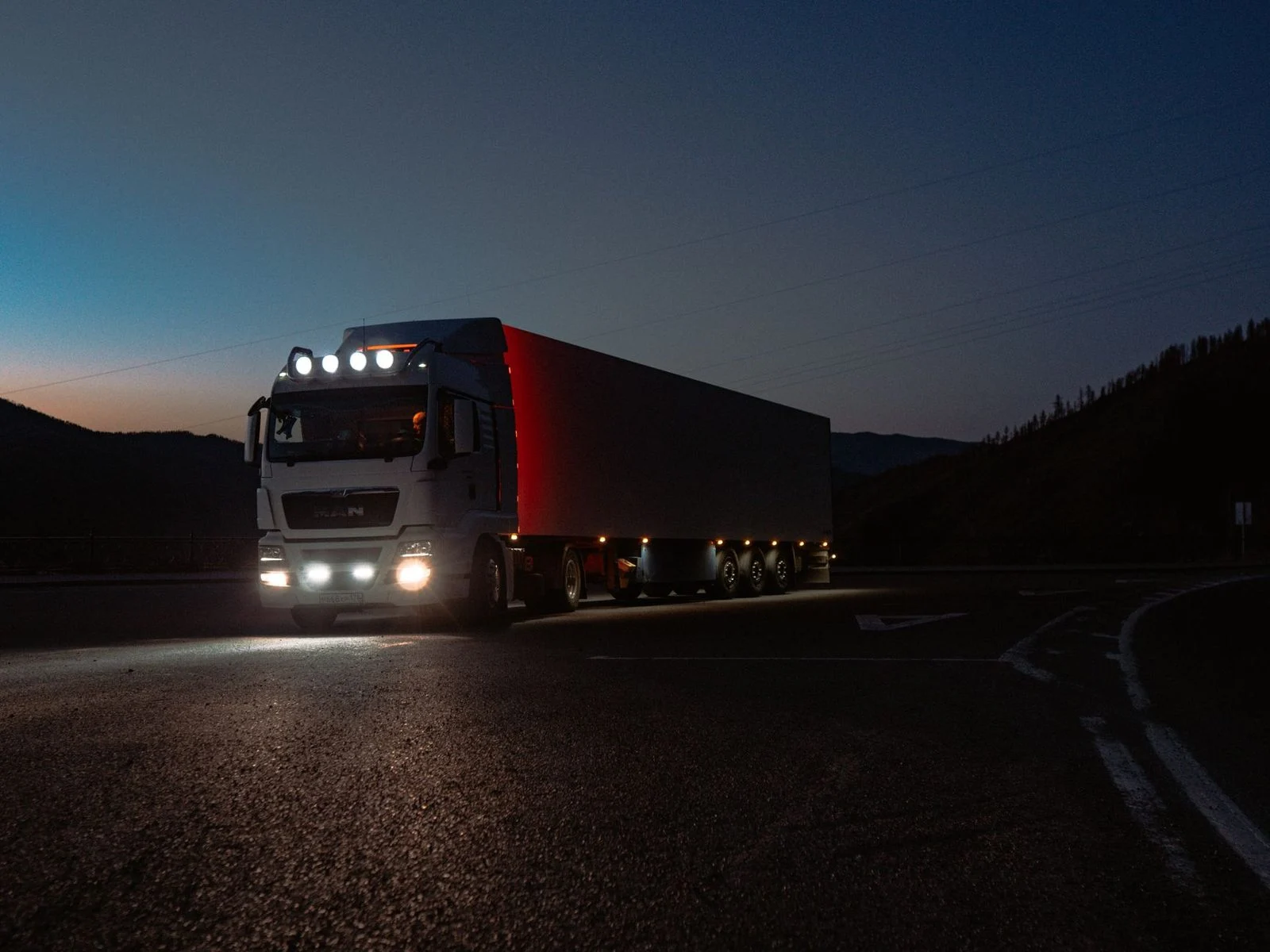 A heavy-duty truck drives along a quiet mountain road at dusk, its headlights lighting the path ahead.