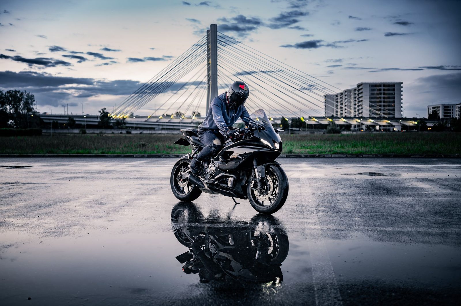A motorcyclist rides a sports bike on a wet road at dusk, with a bridge in the background.