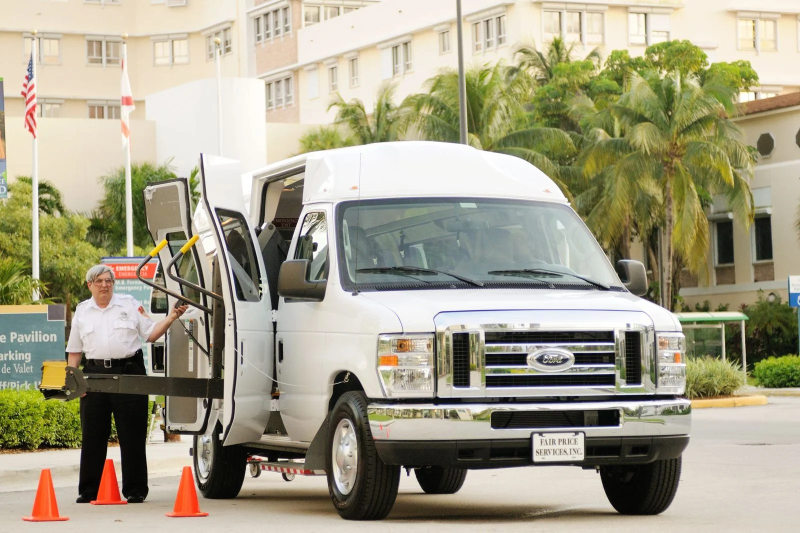 A parking attendant helps a wheelchair user board an accessible van with a rear lift in a palm-lined lot.