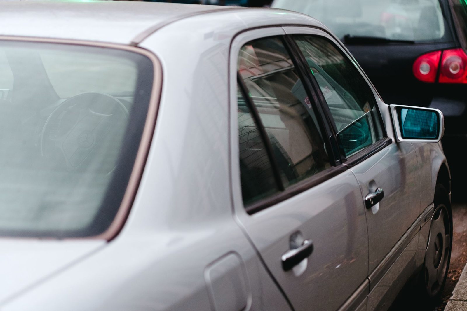 A silver car parked on a city street, with its reflection visible in the side window.