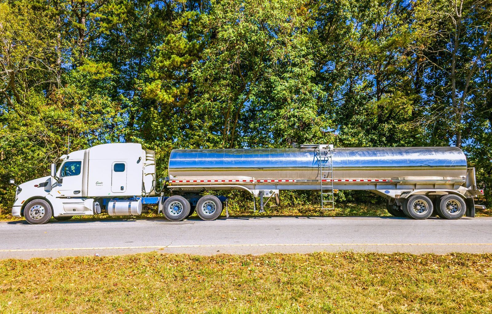 A white semi-truck with a polished tanker trailer parked on a forested roadside during the day.