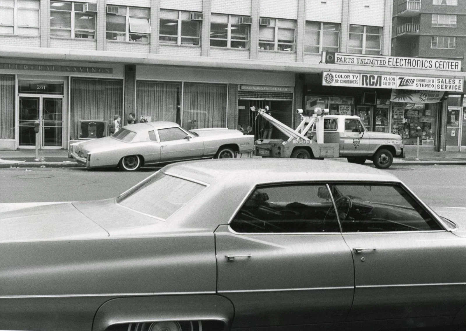 Black-and-white photo of vintage cars and a tow truck on an urban street.