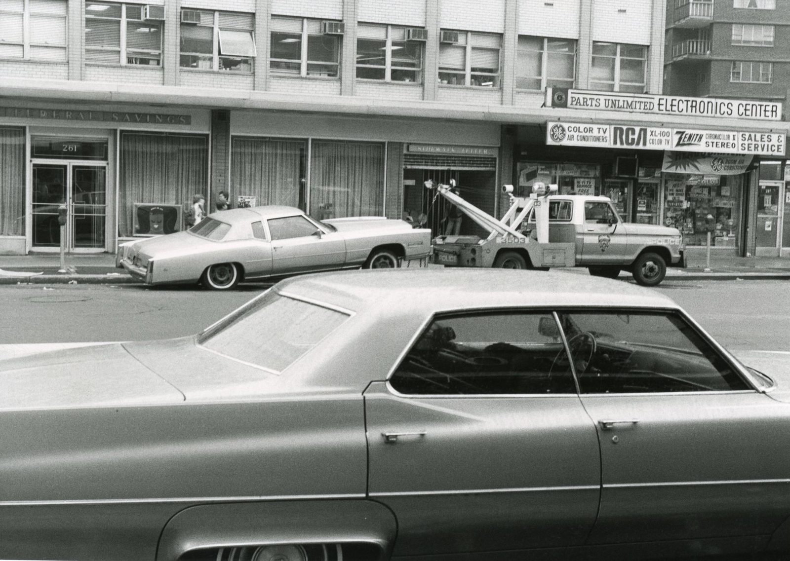 Black-and-white photo of vintage cars and a tow truck on an urban street.