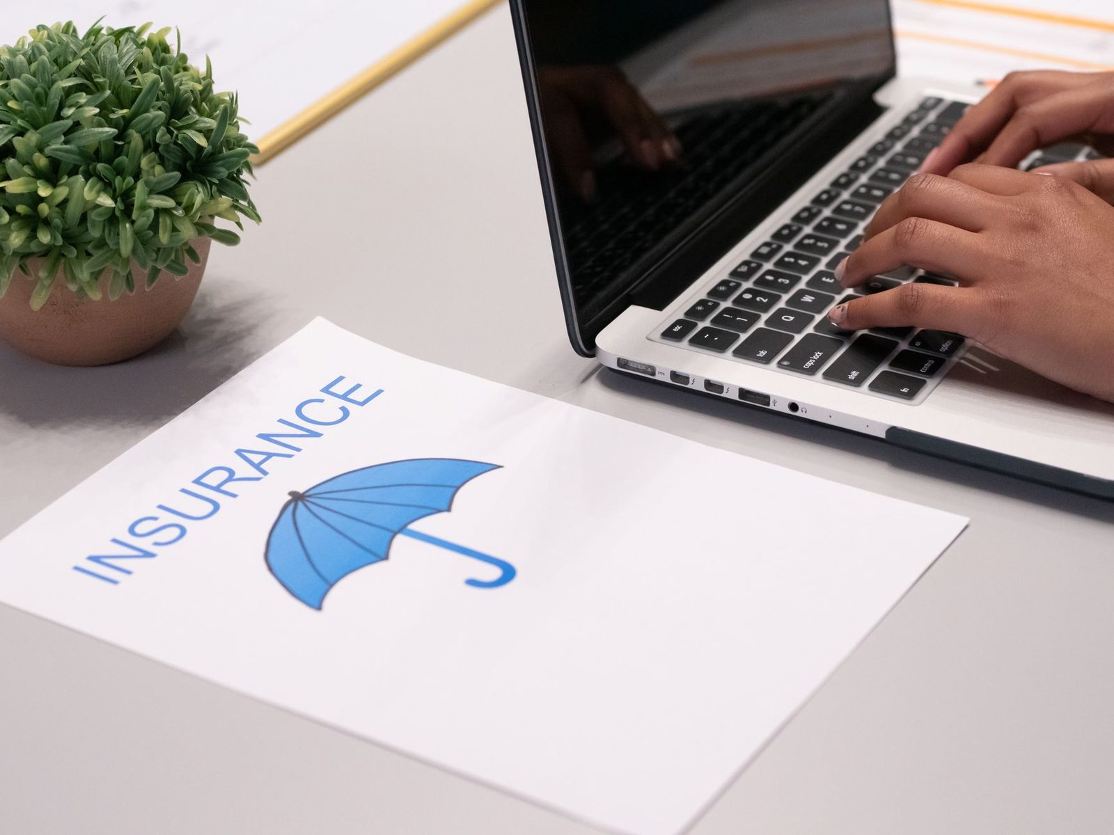 Close-up of hands typing on a laptop beside an insurance document on a desk.