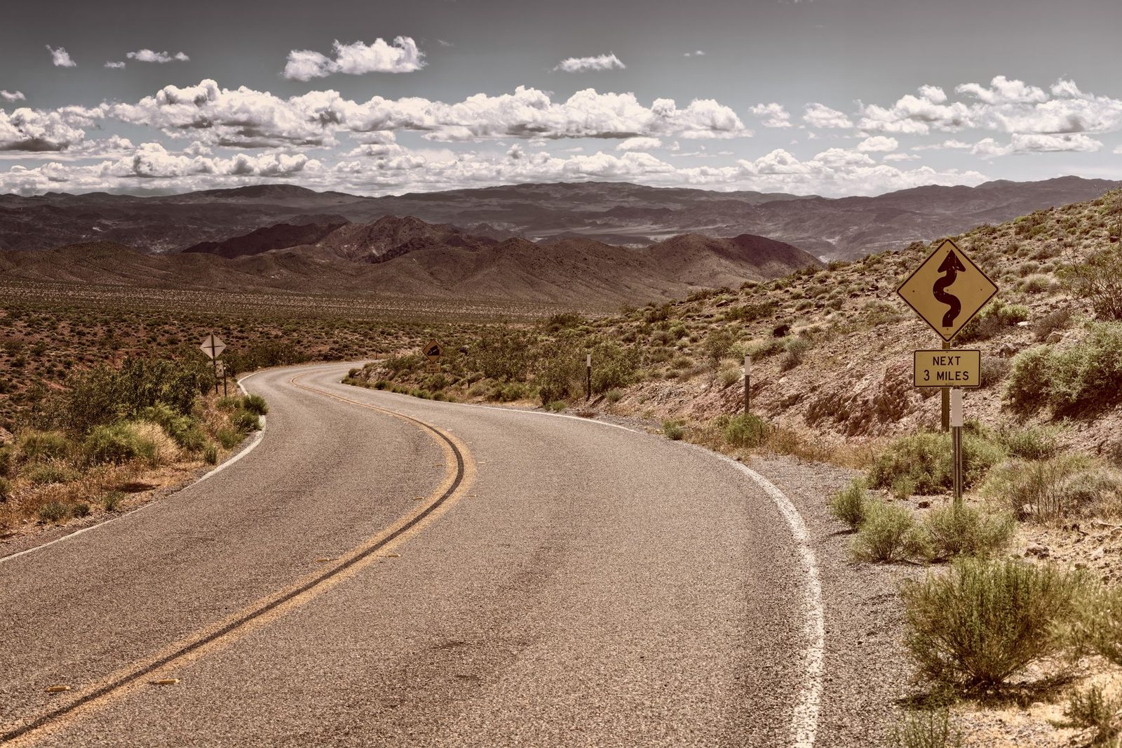 Curved two-lane road winding through an arid desert landscape toward mountains under a clear blue sky.