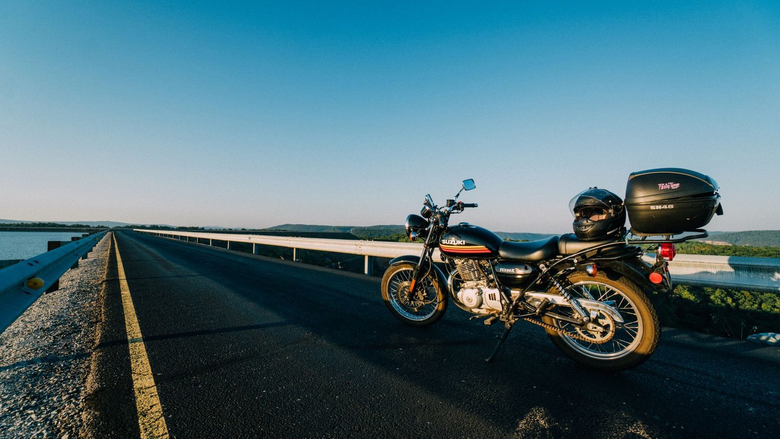 Motorcycle parked on an open highway beside calm water at sunset