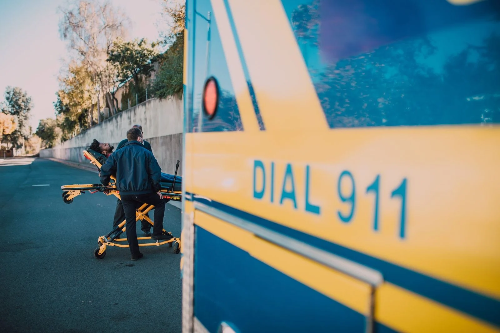 Paramedics wheel a patient on a stretcher beside an ambulance on a sunny day.