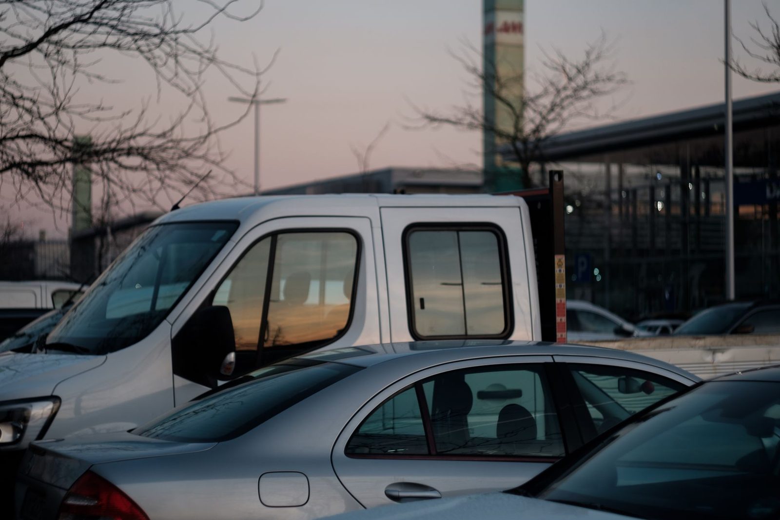 Parked cars line a city street at twilight, with buildings and streetlights in the background.