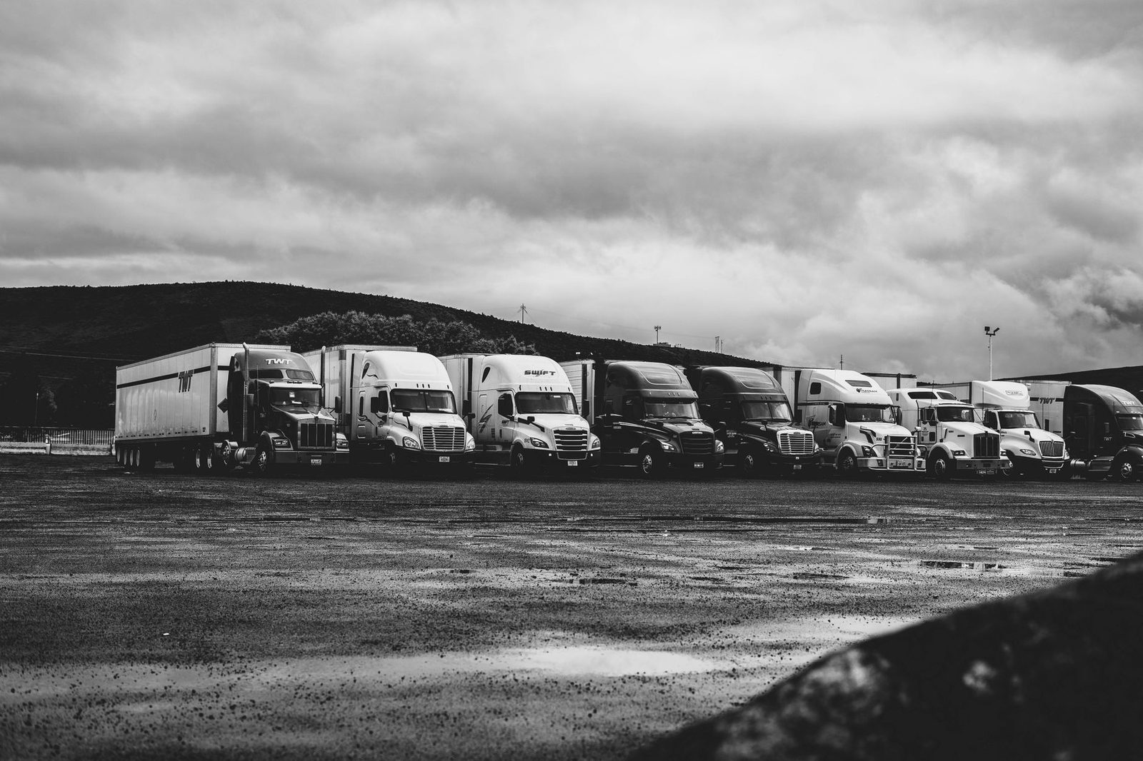 Rows of parked semi trucks sit in a rainy lot in black and white.