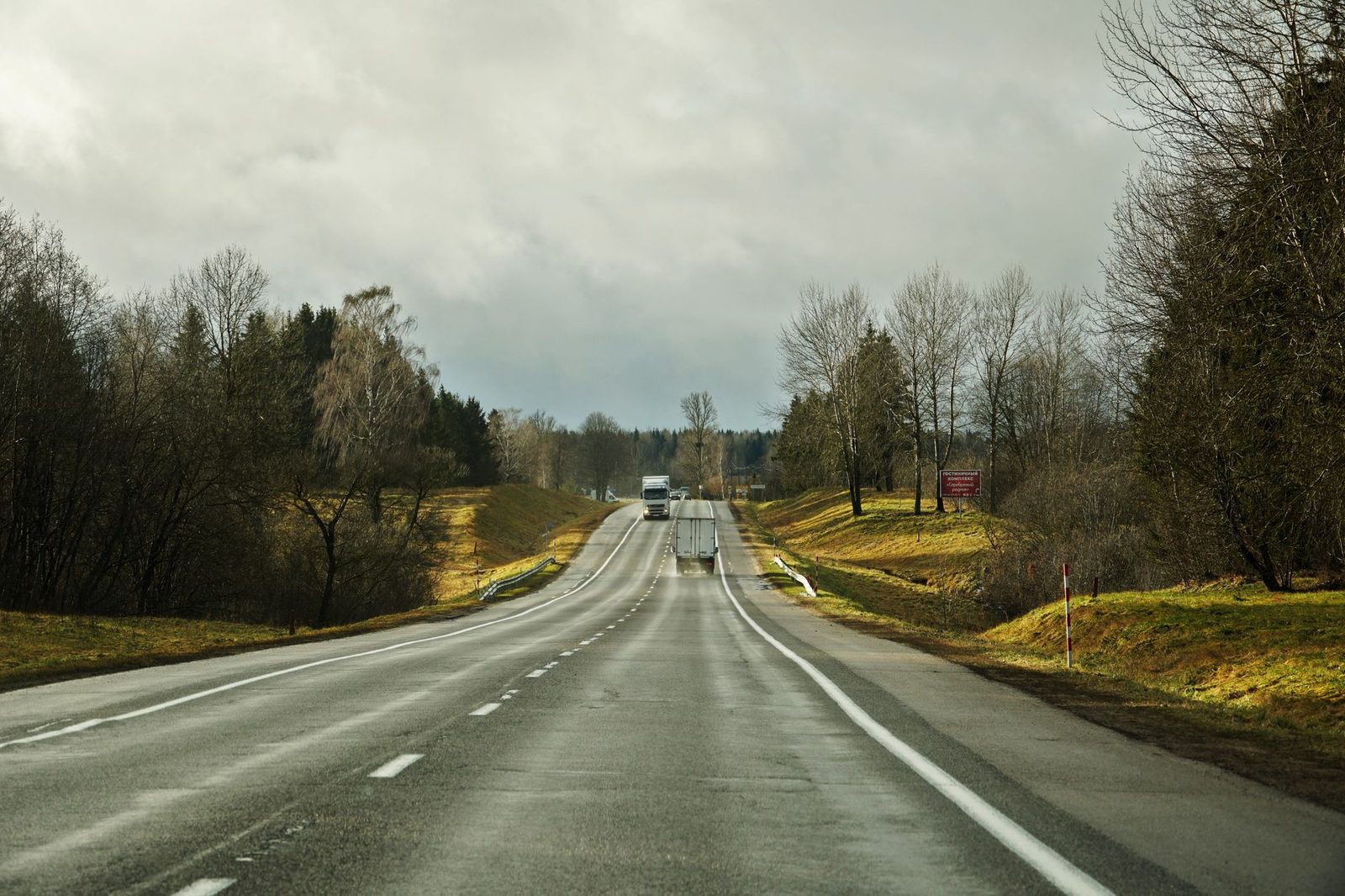 Semi-trucks travel a straight highway under overcast skies, with flat fields on either side.