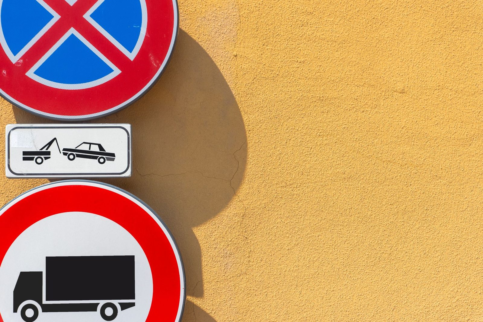 Traffic signs against a bright yellow wall, showing no-parking and no-trucks restrictions.