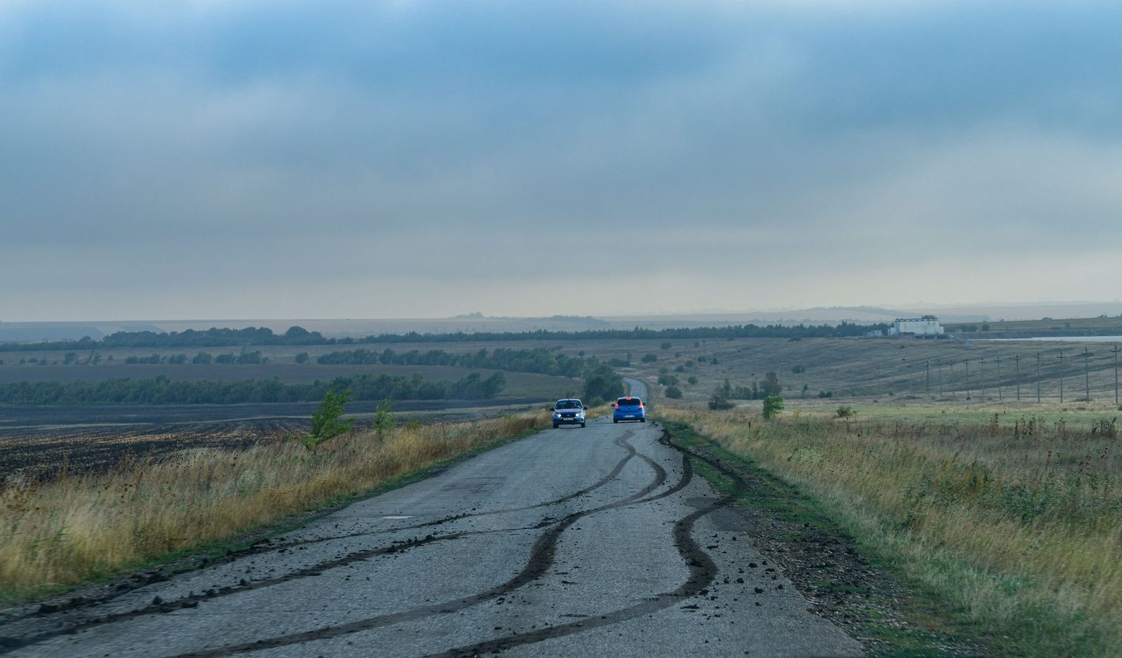 Two cars sit on a rural road with skid marks, with fields and a cloudy sky behind them.