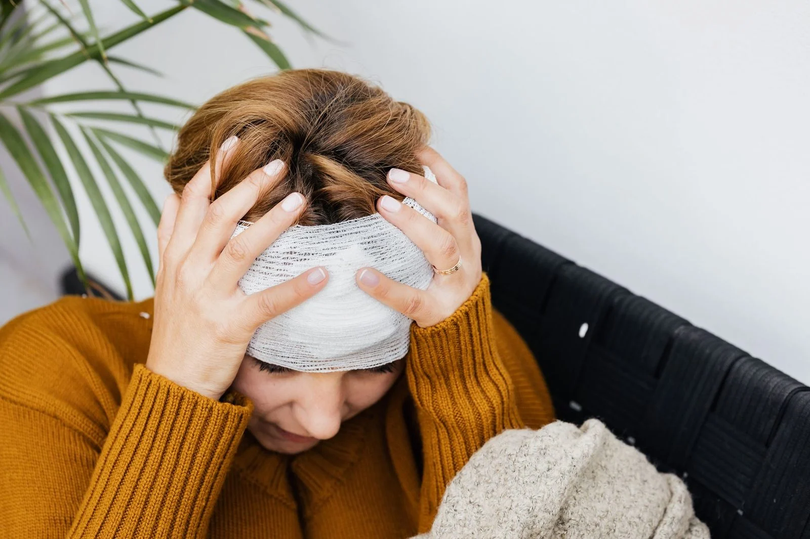 A woman sits indoors holding her head, appearing to be in pain.