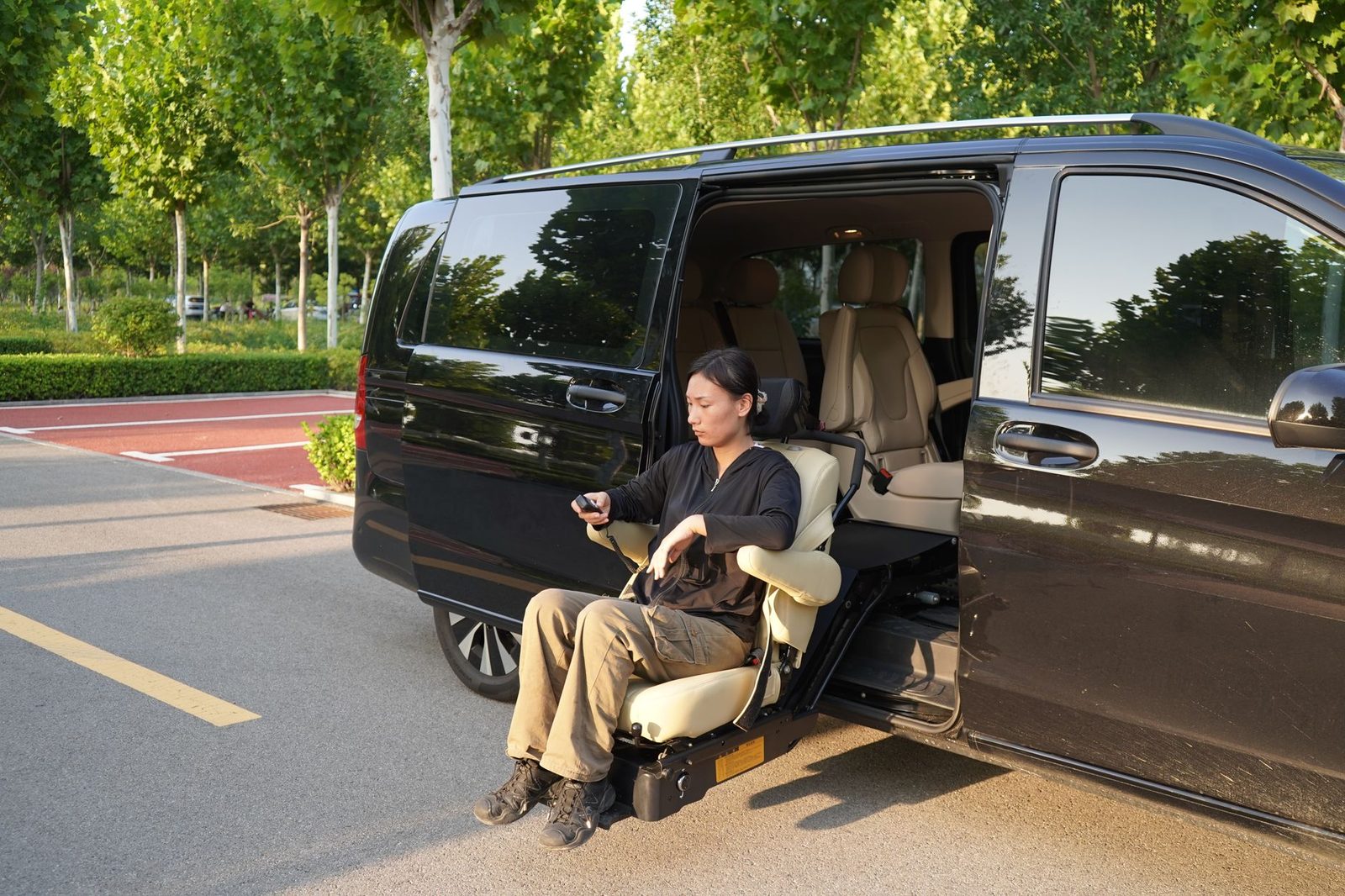 Woman uses a seat lift to enter a black van in a parking lot with trees behind her.